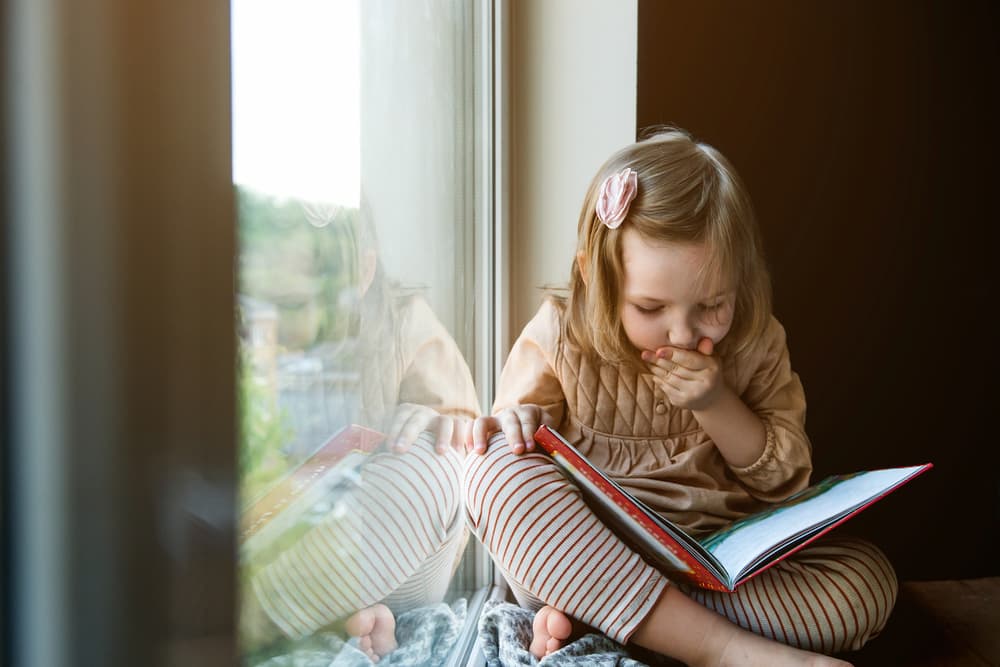 Little girl reading a book by a windowsill